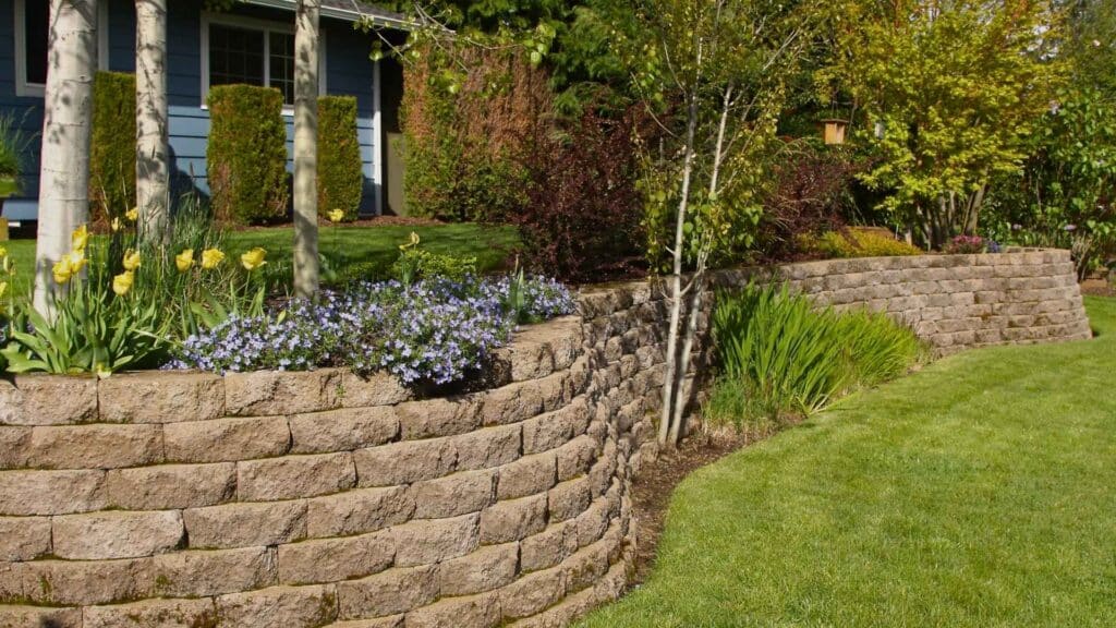 landscaped backyard with stone brick retaining wall with plants and bushes planted above grass